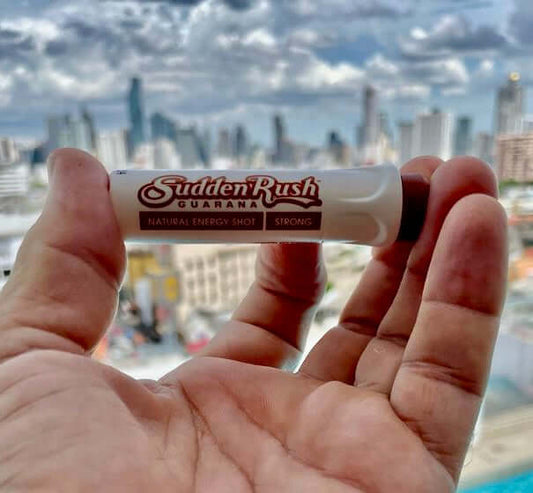 A hand holding a SuddenRush Guarana shot with the Bangkok skyline in the background