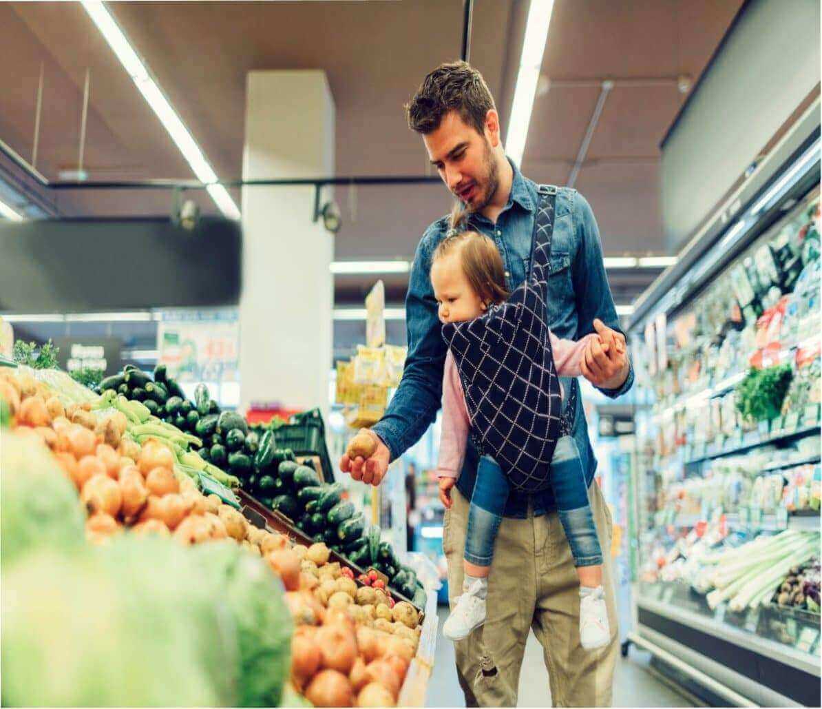 Father grocery shopping with baby in a carrier, selecting fresh produce in a supermarket